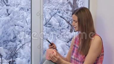 漂亮的女孩坐在窗台上，用智能手机喝茶。 冬天在外面。
